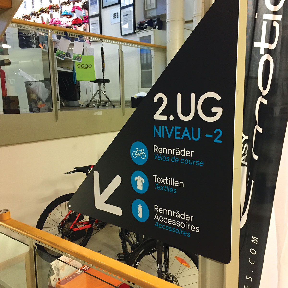 Affichage mural in situ de signalétique triangulaire magasin de vélos, avec indication des produits disponibles à l'étage inférieur.