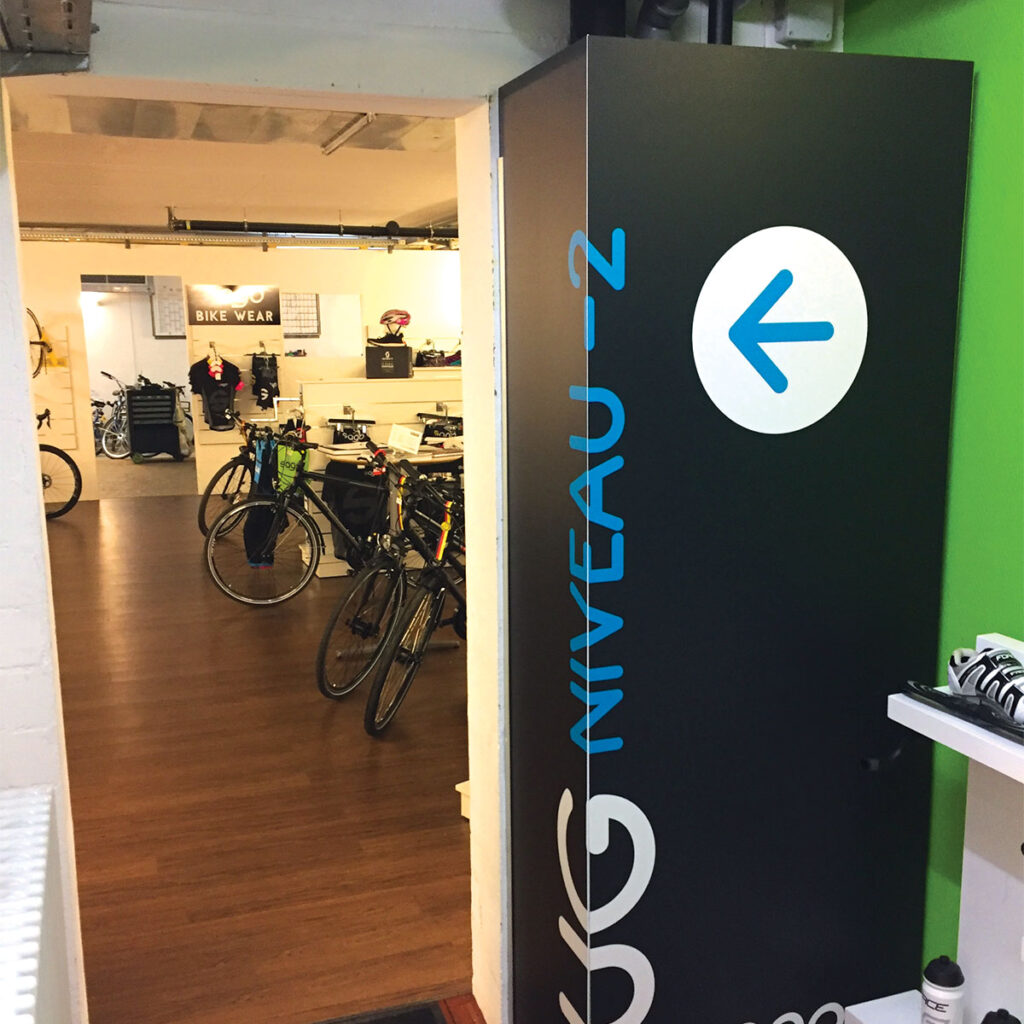 Signalétique magasin de vélos in situ : panneau noir à l’entrée de l’espace vélos de course, marqué "Niveau -2" avec une flèche directionnelle.